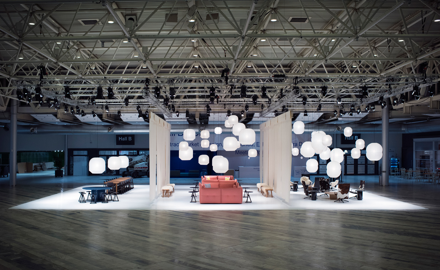 A modern exhibition space with stylish furniture, warm lighting, and hanging spherical lamps, set in a large convention hall. The setup includes sofas, tables, and seating areas arranged symmetrically. - Nolita Harbour
