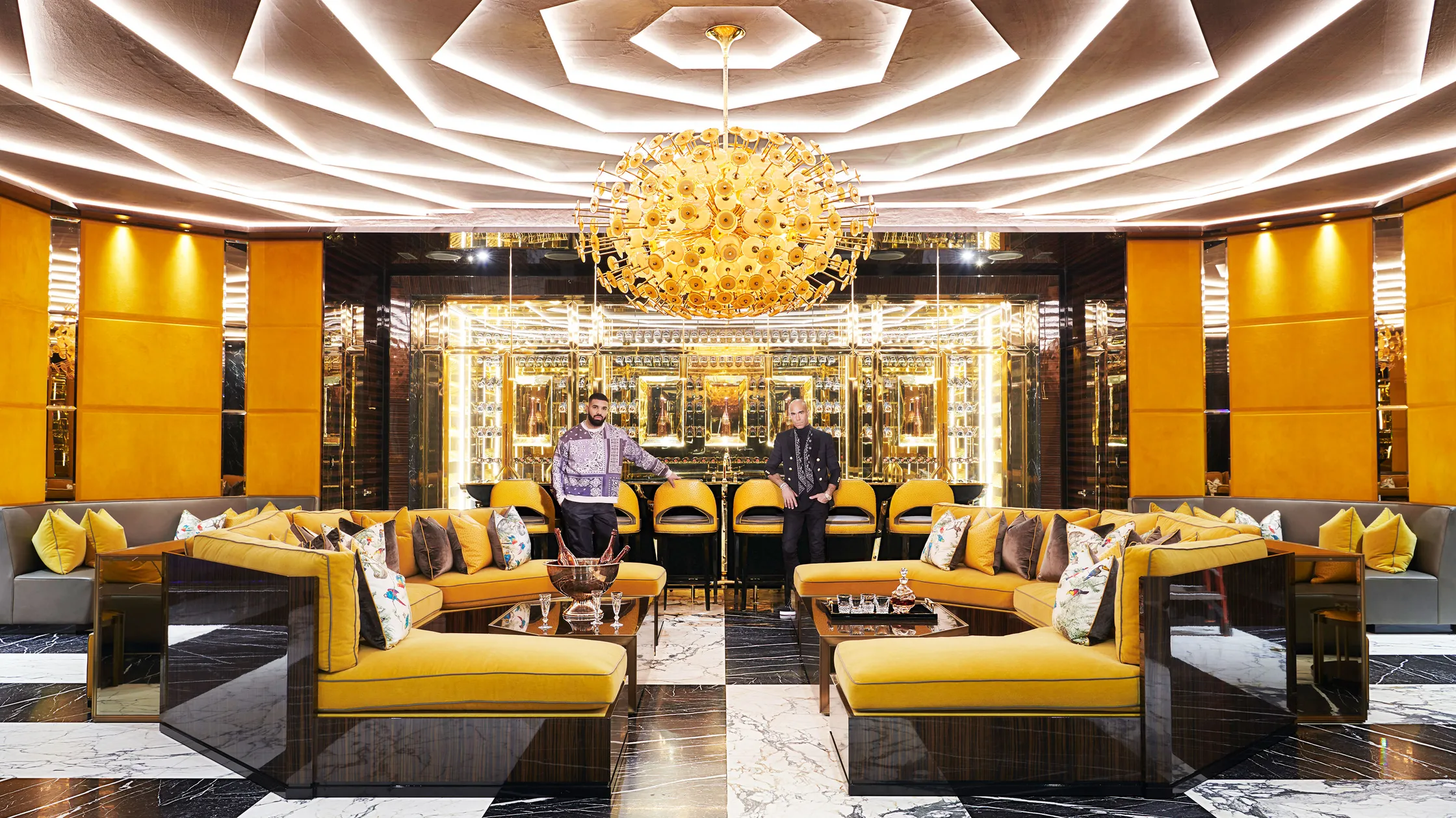 An opulent living space with striking gold and black decor, geometric ceiling lights, mirrored walls, plush yellow sofas, marble flooring, and two men posing in a high-end, extravagant setting. - Nolita Harbour