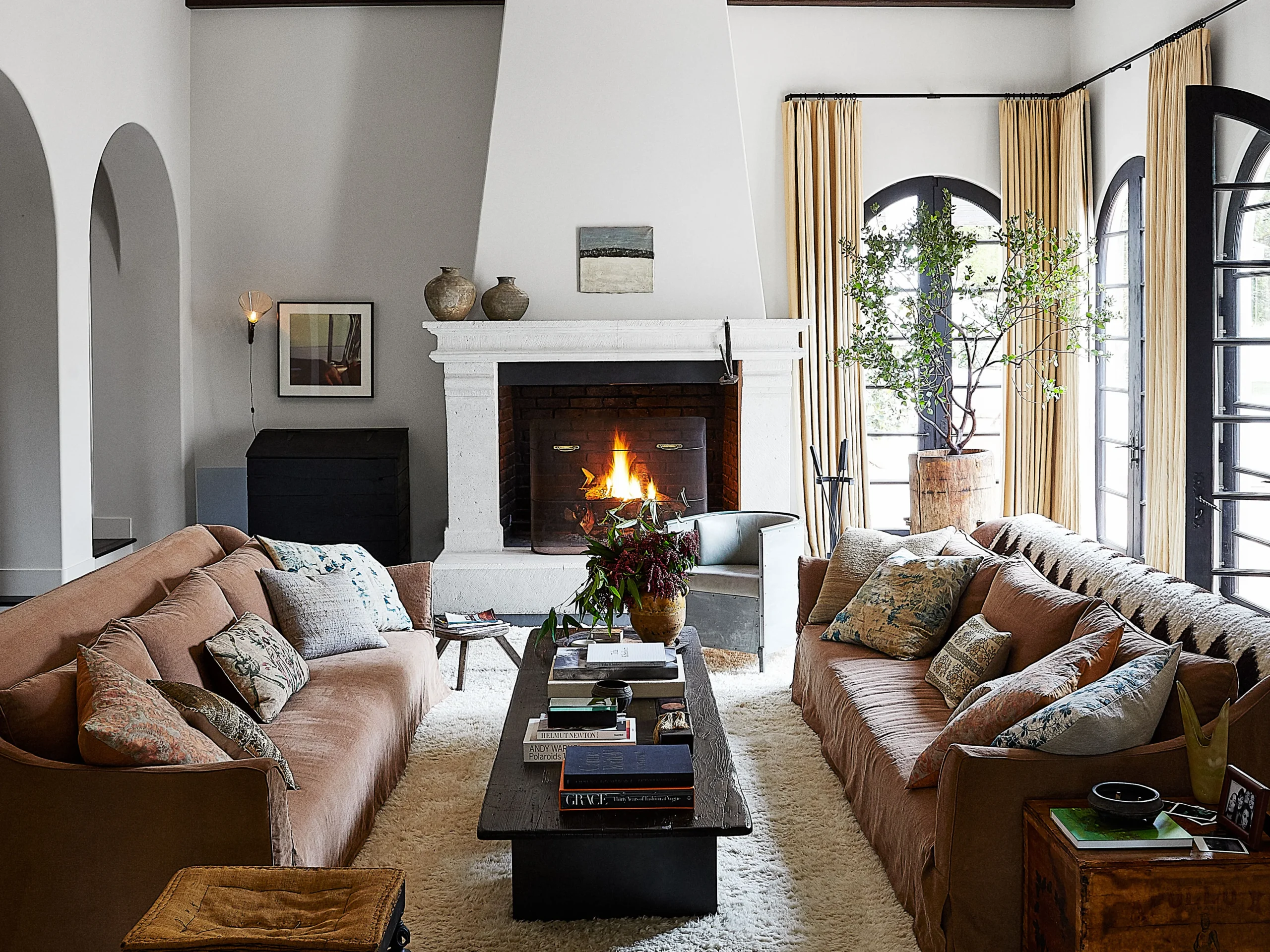 A warm and inviting living room with earthy-toned sofas, a lit fireplace, plush pillows, wooden furniture, and arched windows with flowing curtains, exuding a cozy, vintage charm. - Nolita Harbour