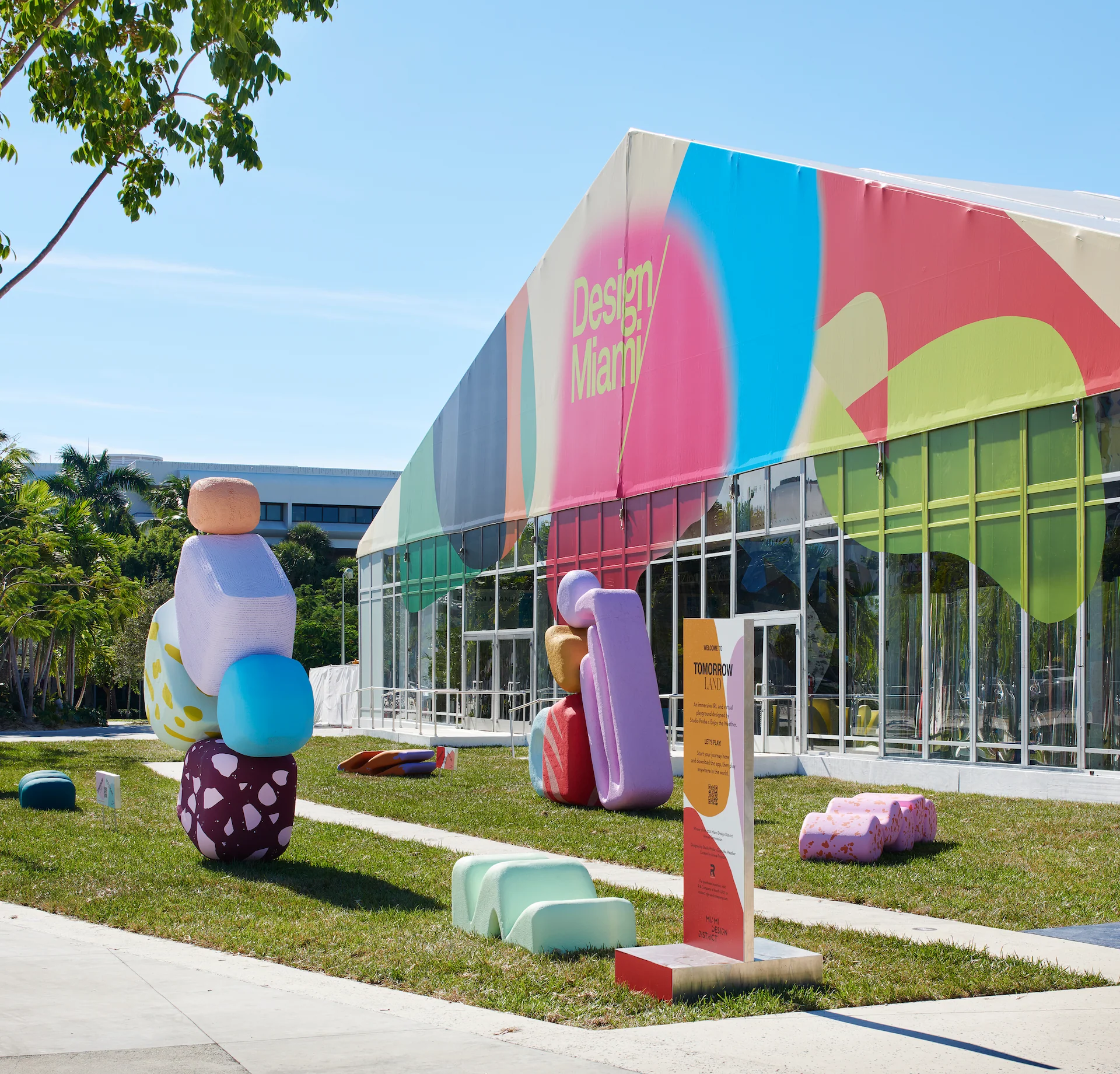 An outdoor scene from Design Miami, a contemporary design fair. A colorful event tent with "Design Miami" branding is visible, along with abstract, oversized sculptural seating elements on a grassy lawn. - Nolita Harbour