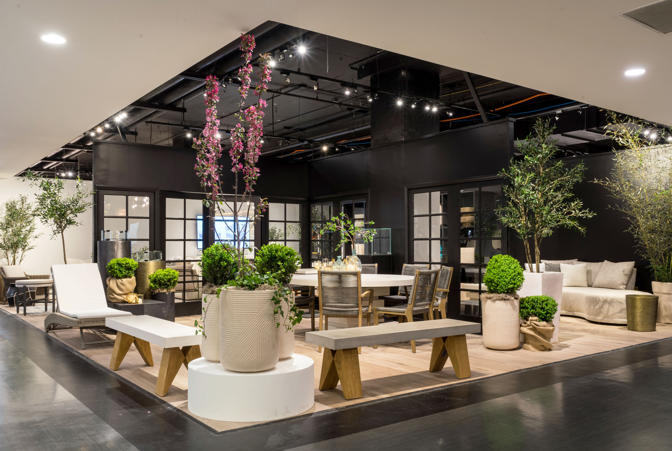 A modern interior design showroom featuring contemporary outdoor furniture, natural wood benches, and woven planters with lush greenery. The space has a neutral color palette with black-framed glass partitions, soft lighting, and a stylish mix of textures, creating an elegant and inviting atmosphere. - Nolita Harbour 