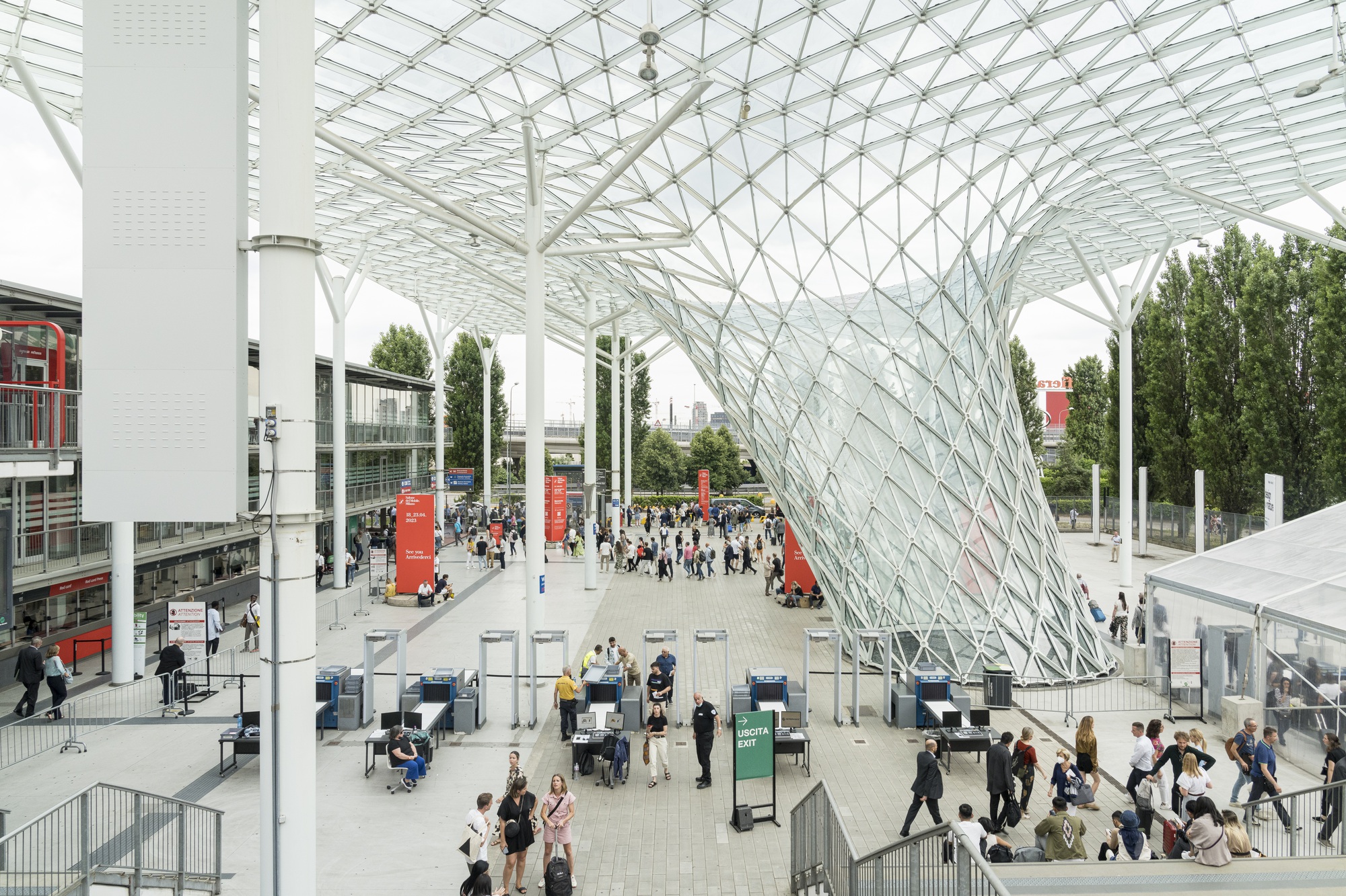 The image showcases a modern exhibition venue with a striking glass and steel structure. Attendees are seen entering and exiting through security checkpoints, while others walk through the spacious outdoor area. The scene reflects a lively and well-organized event, likely an international trade fair or design exhibition. - Nolita Harbour 