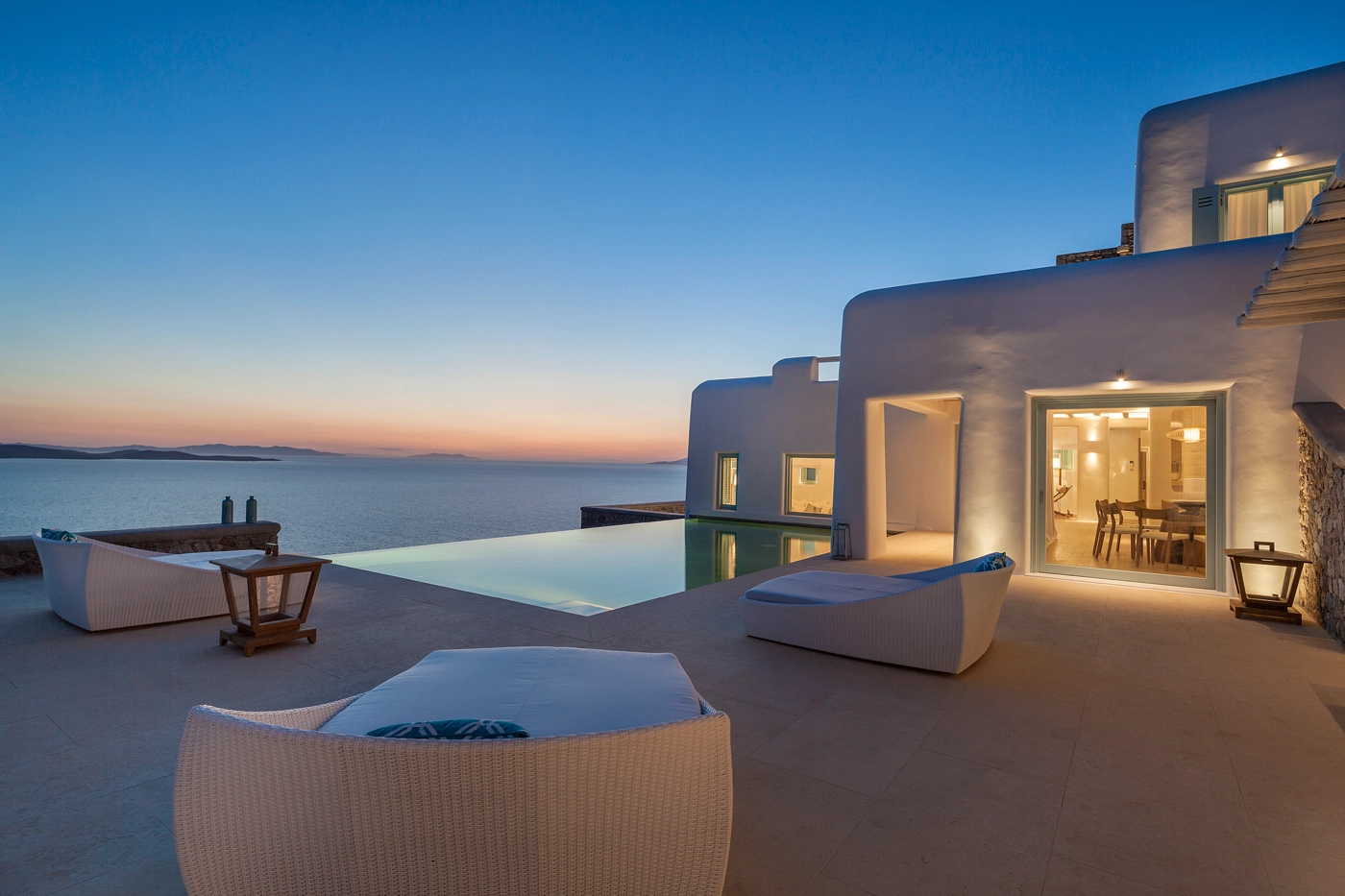 Luxury Villas of Mykonos with infinity pool overlooking the sea at sunset, featuring modern white architecture and outdoor lounge seating.”
