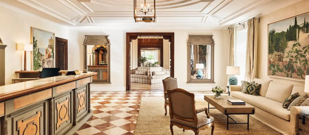 Elegant Mediterranean-style hotel reception with checkered marble floors, classic wooden furniture, and a cozy lounge area with beige sofa and armchairs.