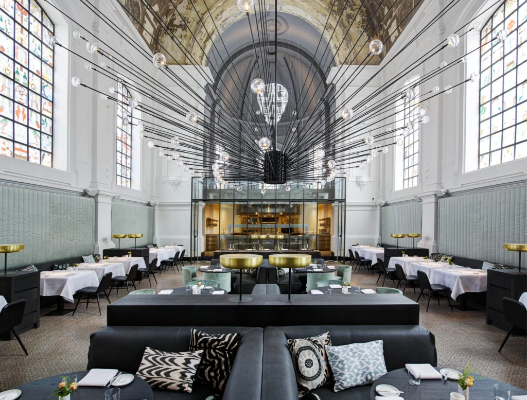 Grand luxury restaurant interior set inside a restored historic hall, featuring a dramatic sculptural chandelier, soaring vaulted ceilings, elegant table settings, and contemporary fine dining design within a heritage architectural setting.