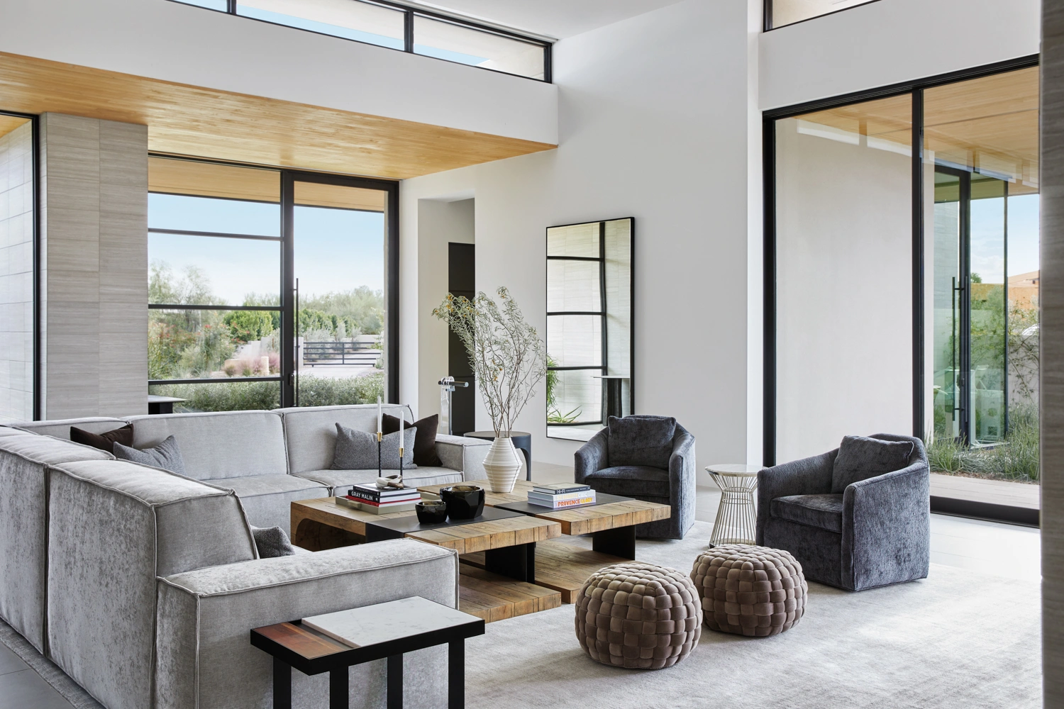 Bright modern living room with a large gray sectional sofa, matching armchairs, wooden coffee tables, woven ottomans, and floor-to-ceiling windows opening to a landscaped view.