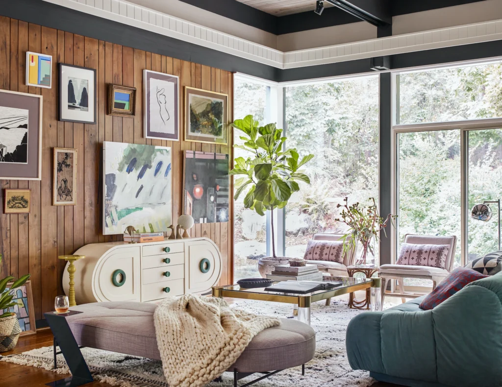 Eclectic living room with wood-paneled walls, a gallery of framed artwork, a sculptural sideboard, plush seating, layered textiles, and large windows overlooking greenery.