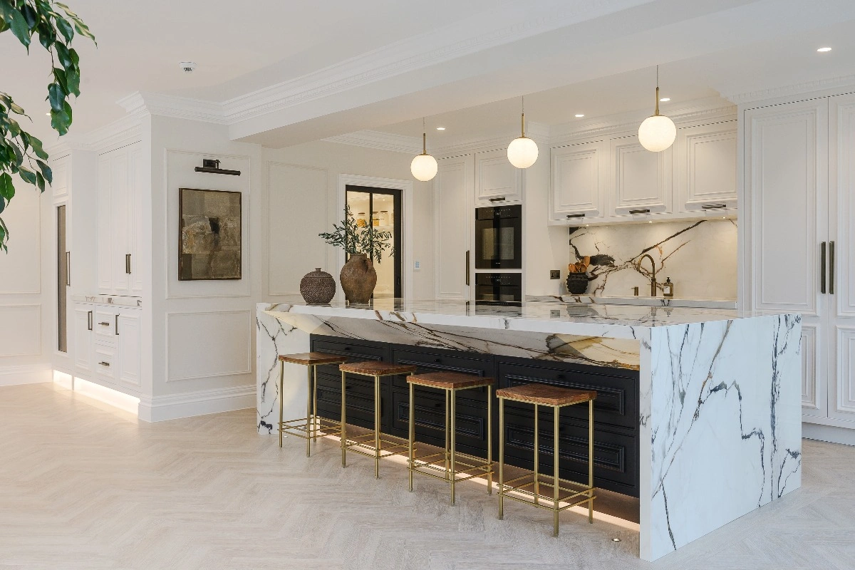 Luxury white kitchen with a marble waterfall island, black cabinetry, gold-accent bar stools, globe pendant lights, and classic panel detailing.