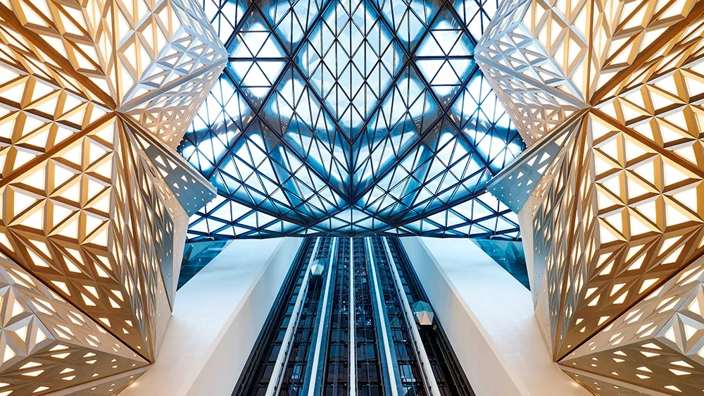 Striking contemporary architectural atrium with geometric metal lattice structures and a dramatic glass ceiling, illustrating the role of architecture as experience in modern luxury design.